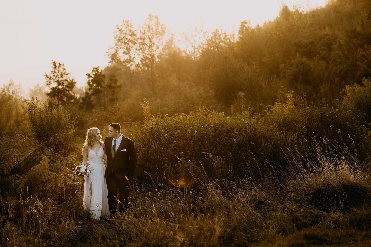 Bride and groom share romantic moment in golden sunset light surrounded by lush green hillside.