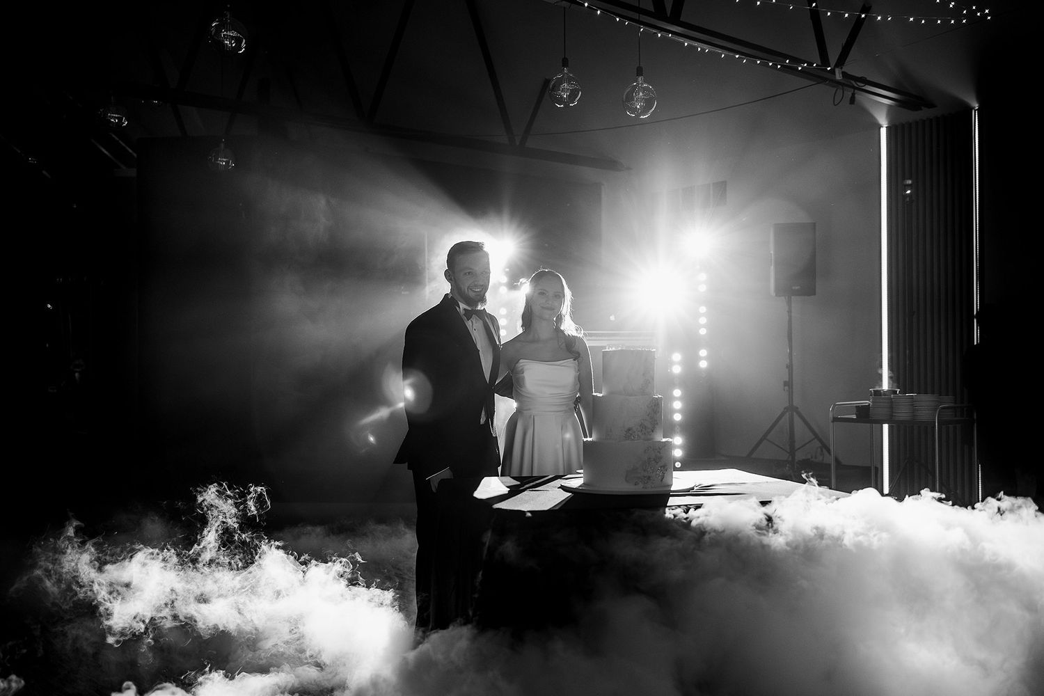 Black and white photo of couple cutting wedding cake surrounded by dramatic fog and stage lighting.