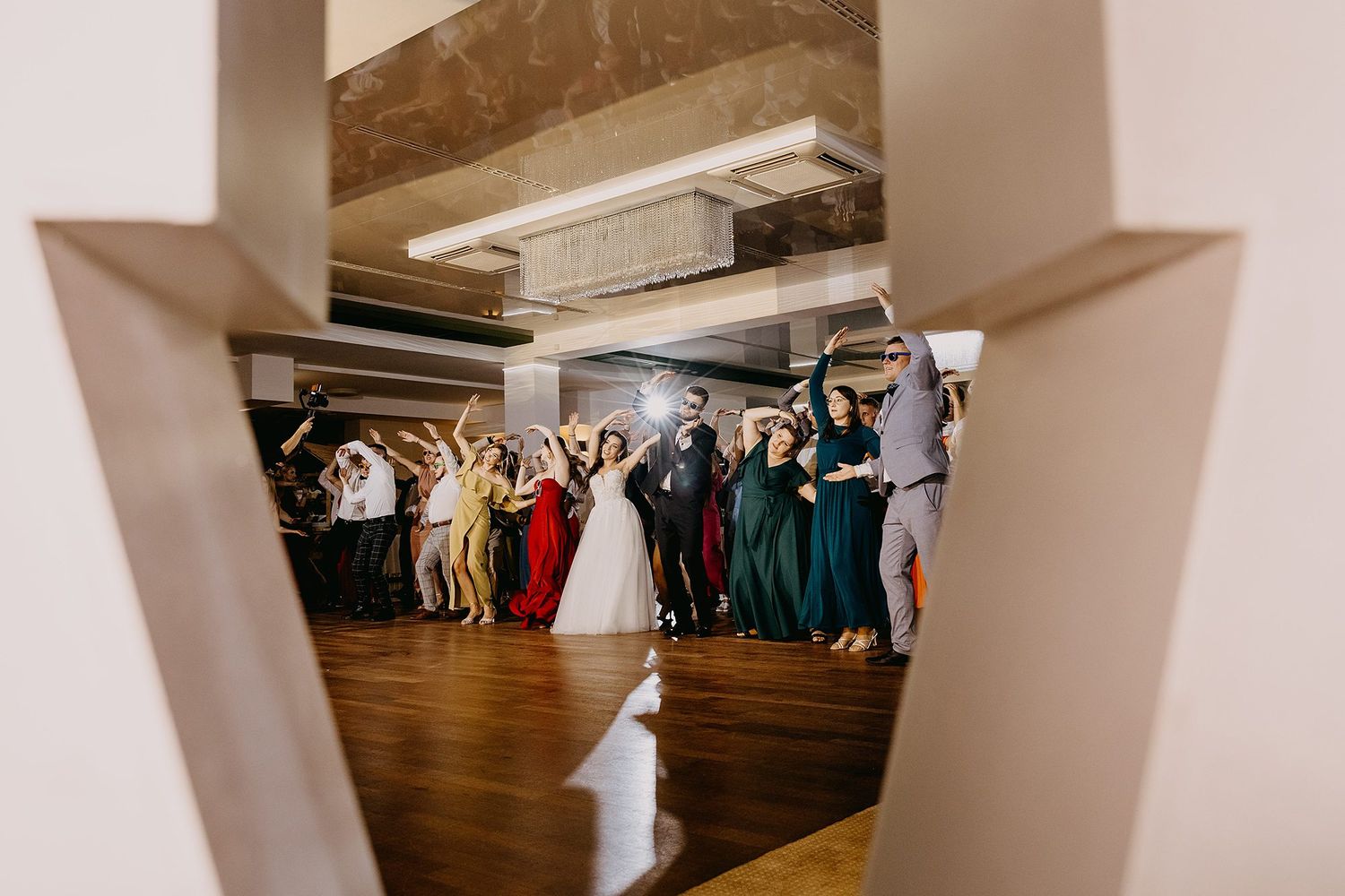 Wedding party celebrating on dance floor, bride in white gown with bridesmaids in emerald dresses, viewed through decorative frame.