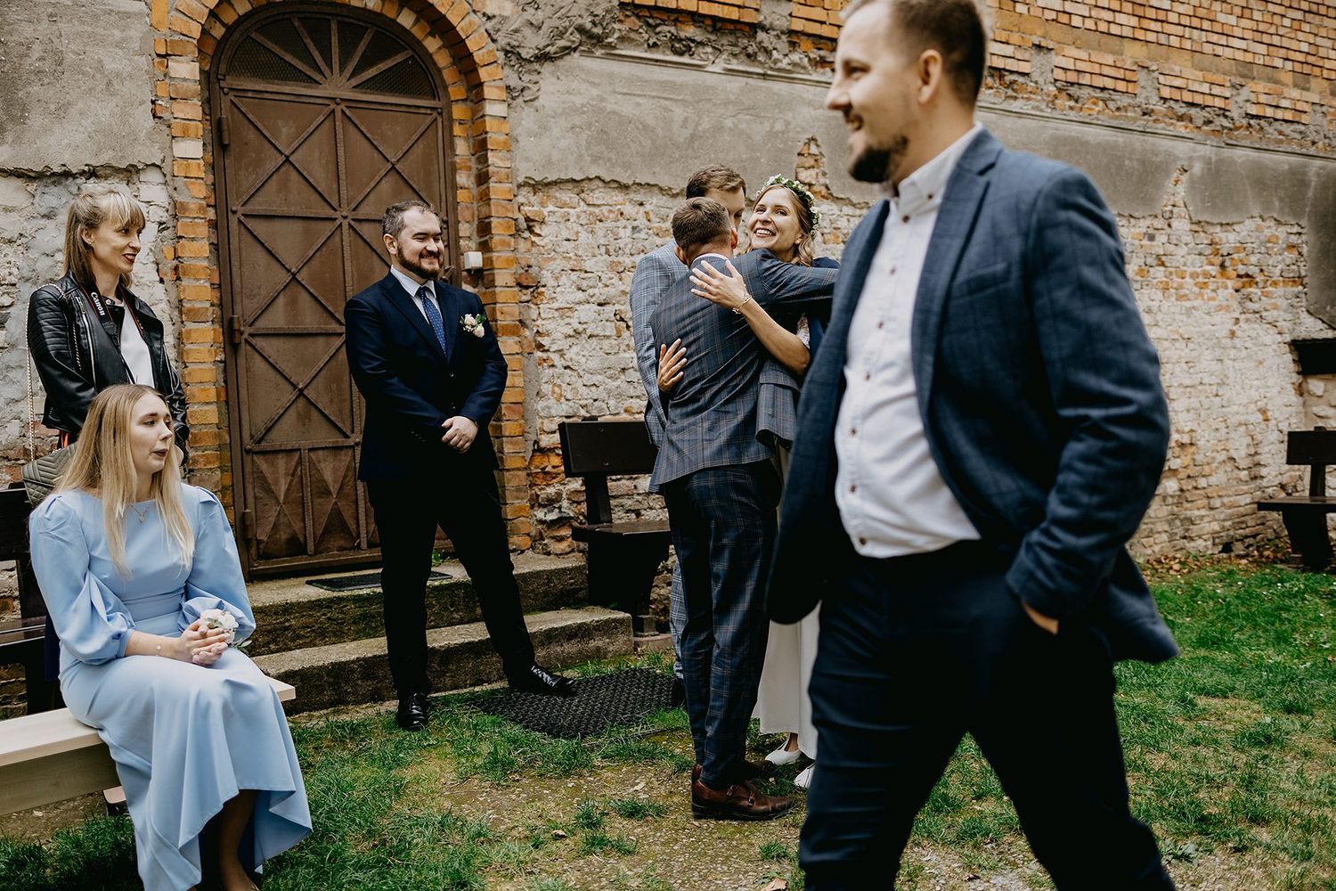 Wedding guests socializing outdoors near rustic brick wall, couple embracing while others watch and chat.