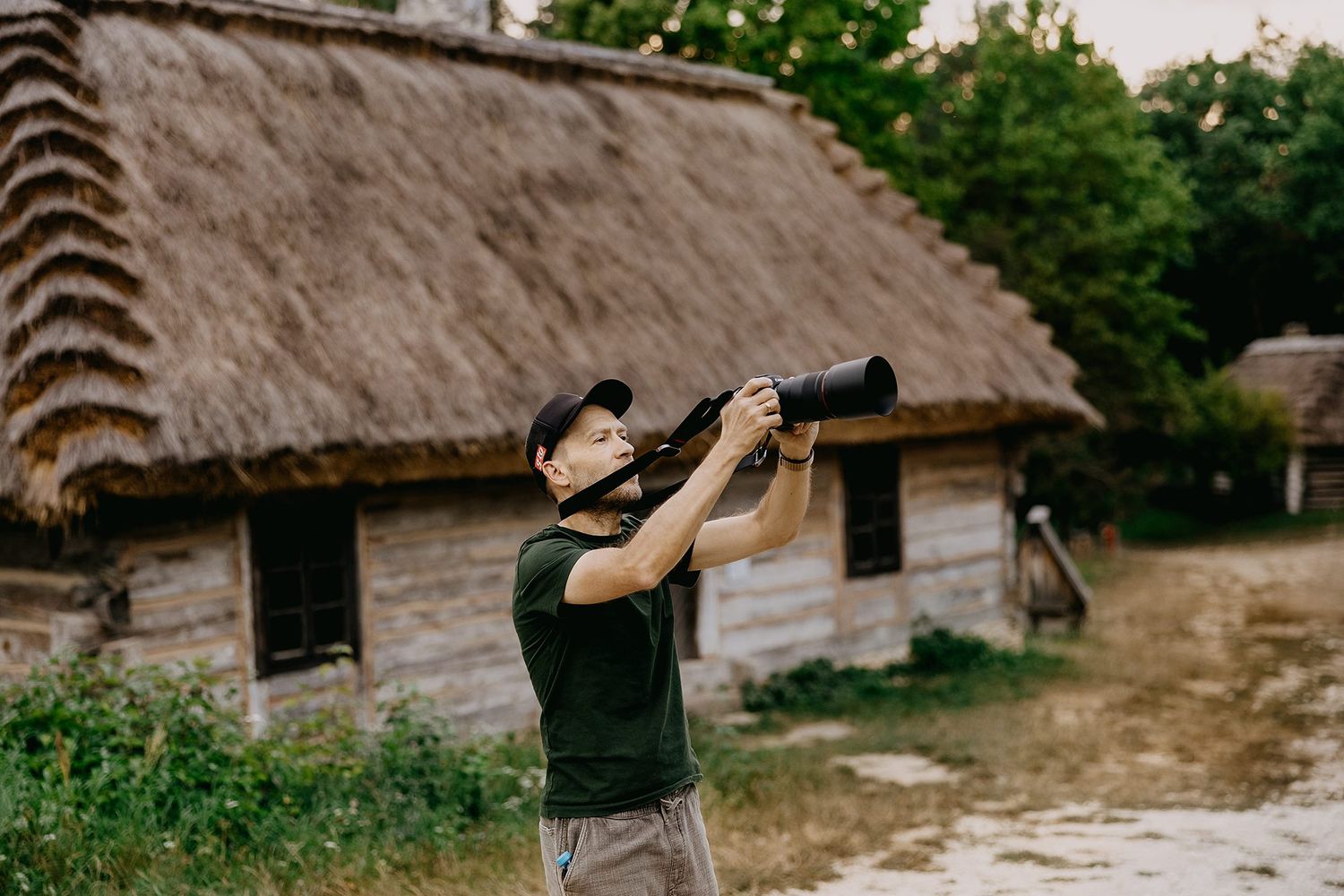 Male photographer using telephoto lens outdoors near rustic thatched-roof wooden countryside building.