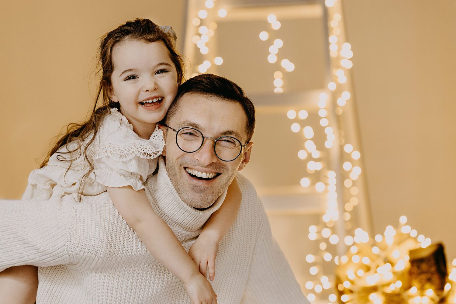 Laughing father giving his cheerful little daughter a piggyback ride near sparkling Christmas lights.