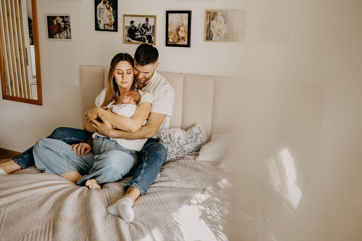 A loving couple sits on a bed together, cradling their newborn baby in warm light.