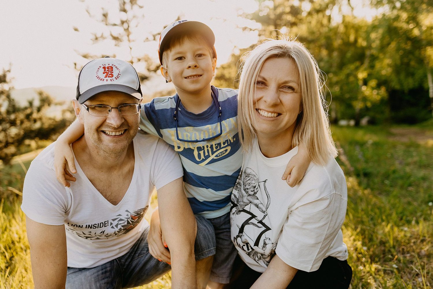 Smiling family of three posing together outdoors in golden sunlight, parents hugging their young son.