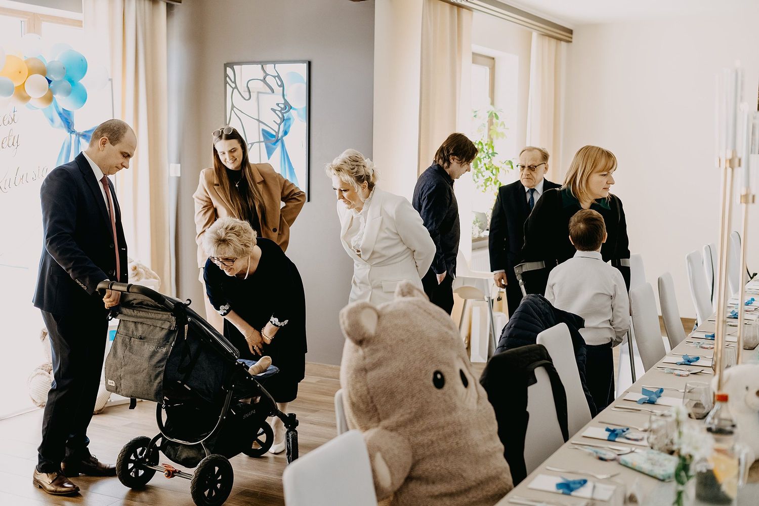 Family members gather in a decorated reception hall for a christening celebration.