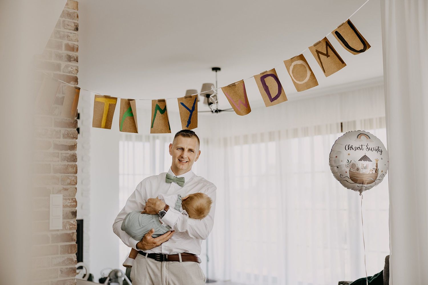 Father in white shirt and bow tie holding baby under festive banner reading TATY DOMU at home celebration.