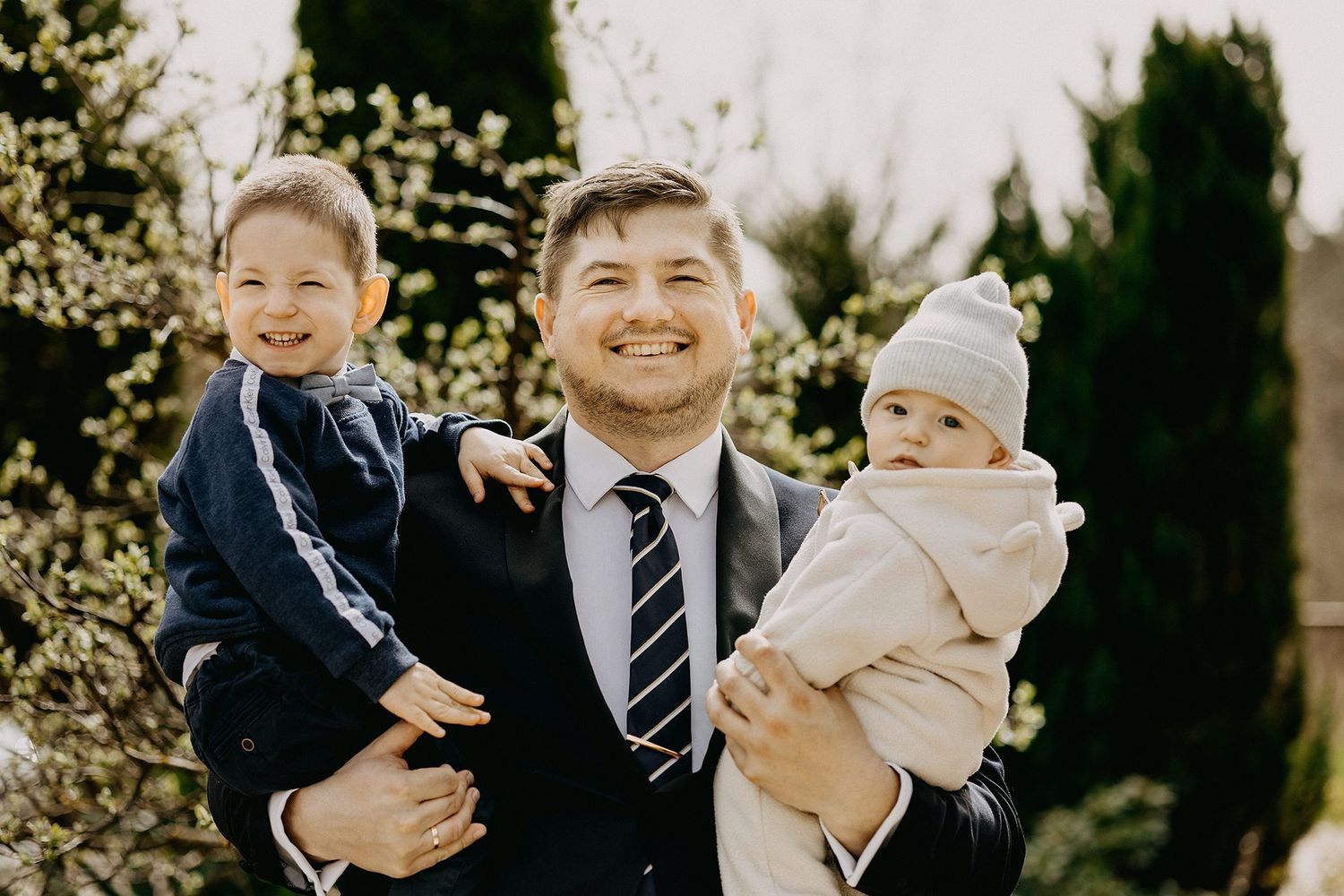 Smiling man in suit holds two young children outdoors near blooming spring trees.