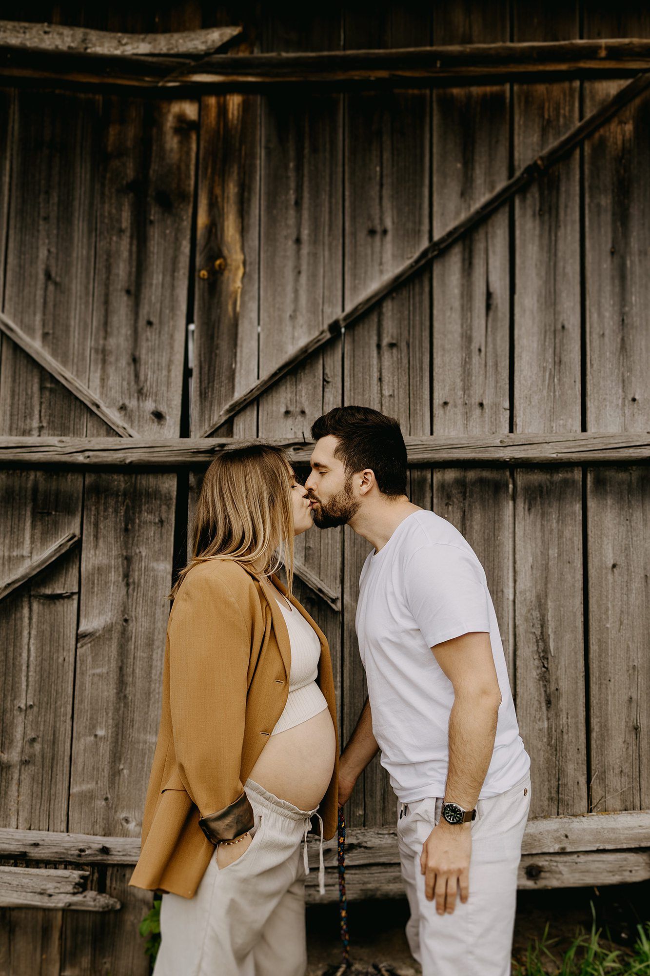 Pregnant couple sharing a kiss in front of rustic wooden barn doors, woman in tan blazer.