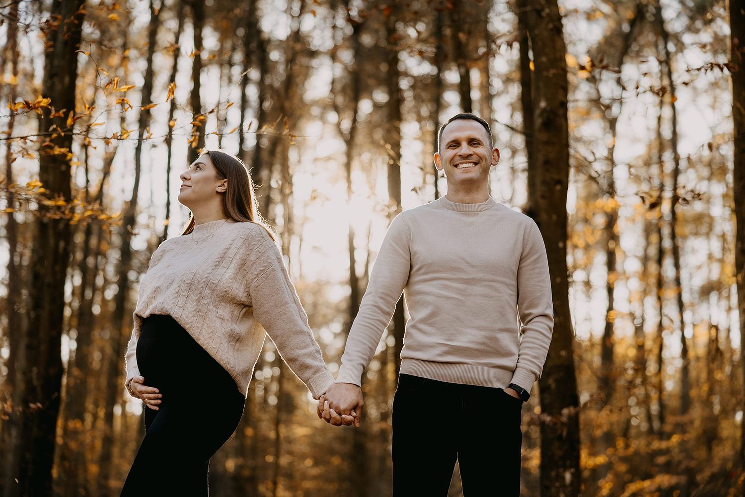 Expectant couple holding hands in golden autumn forest, woman cradling baby bump at sunset.