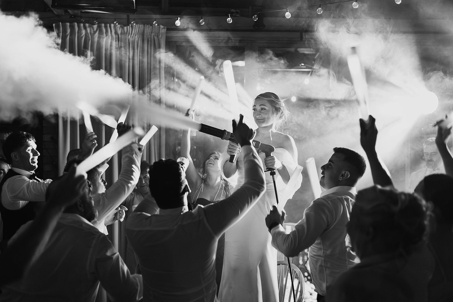 Black and white photo of bride being lifted by guests during a lively wedding celebration with smoke effects.