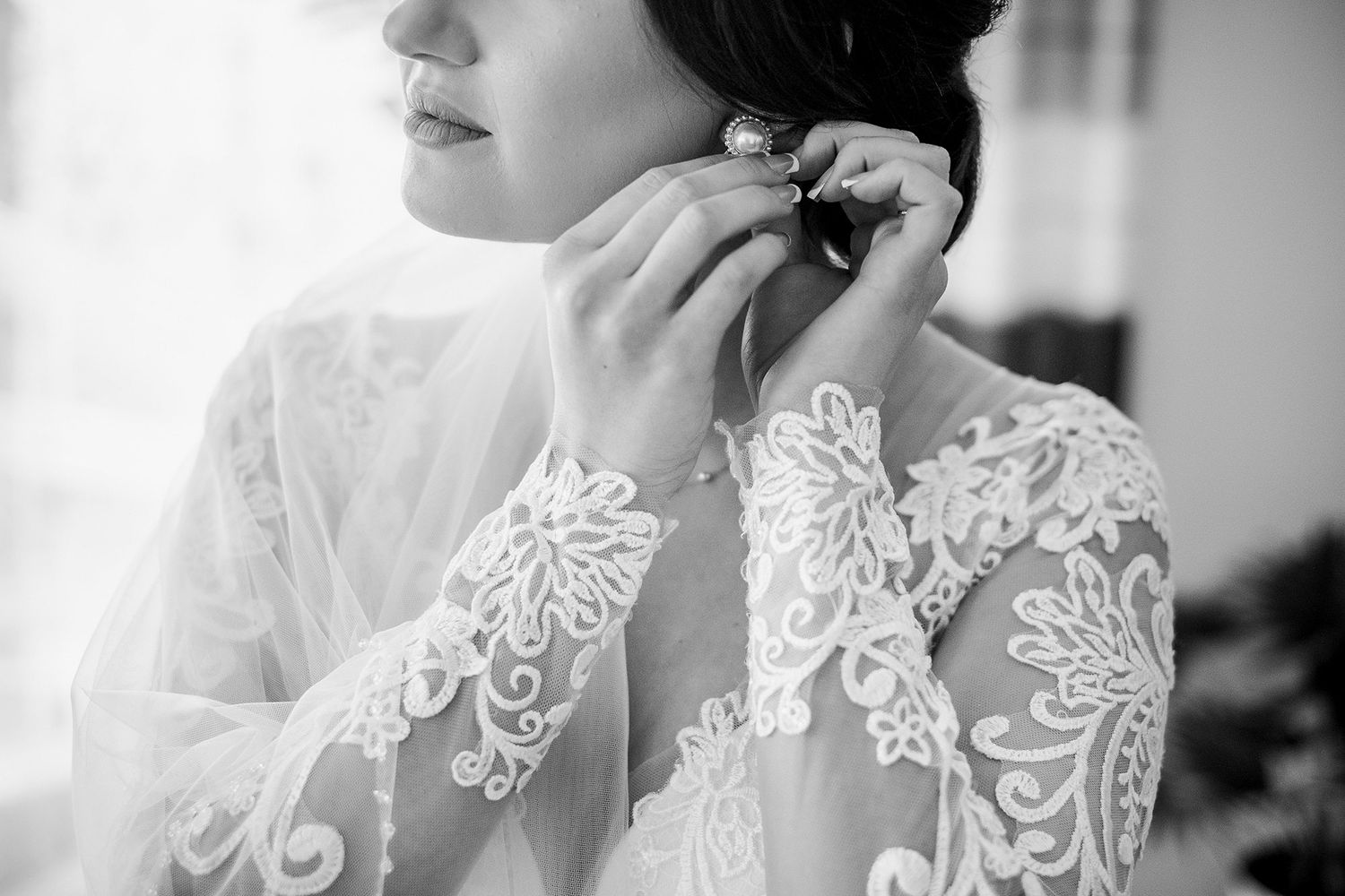 Close-up black and white photo of bride in lace gown putting on pearl earrings before wedding.