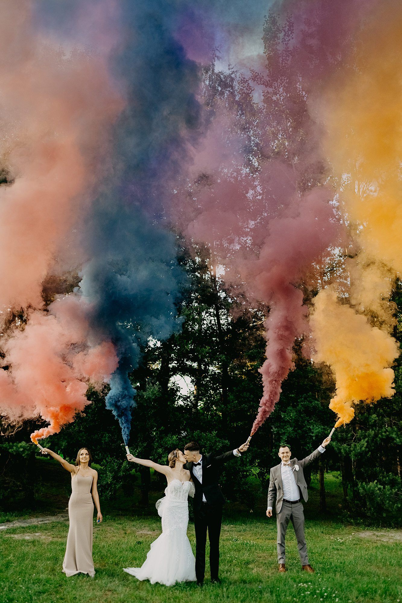 Wedding party holding colorful smoke bombs creating vibrant rainbow clouds outdoors.