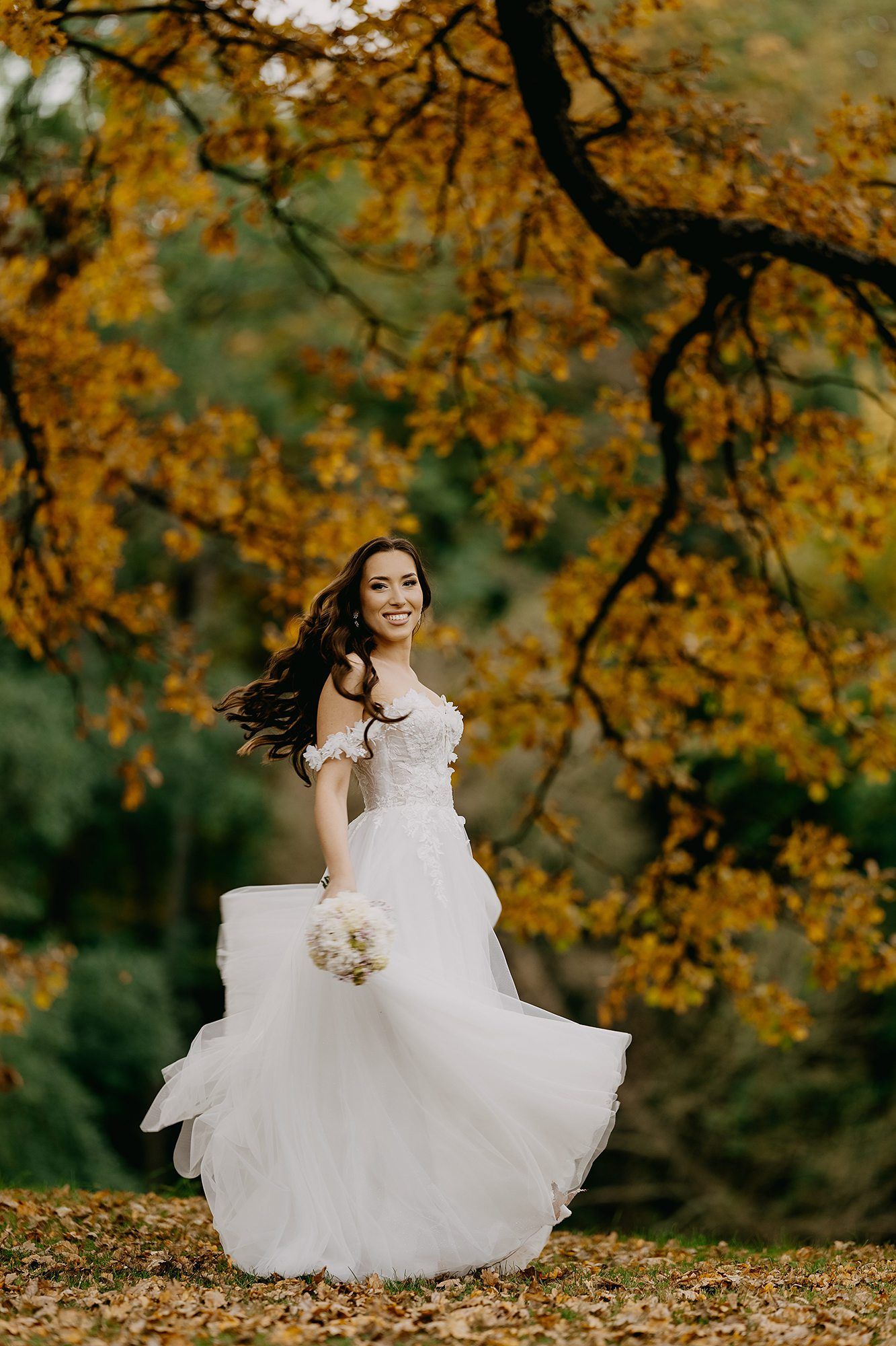 Joyful bride in white off-shoulder gown twirls under golden autumn oak tree in lush park.