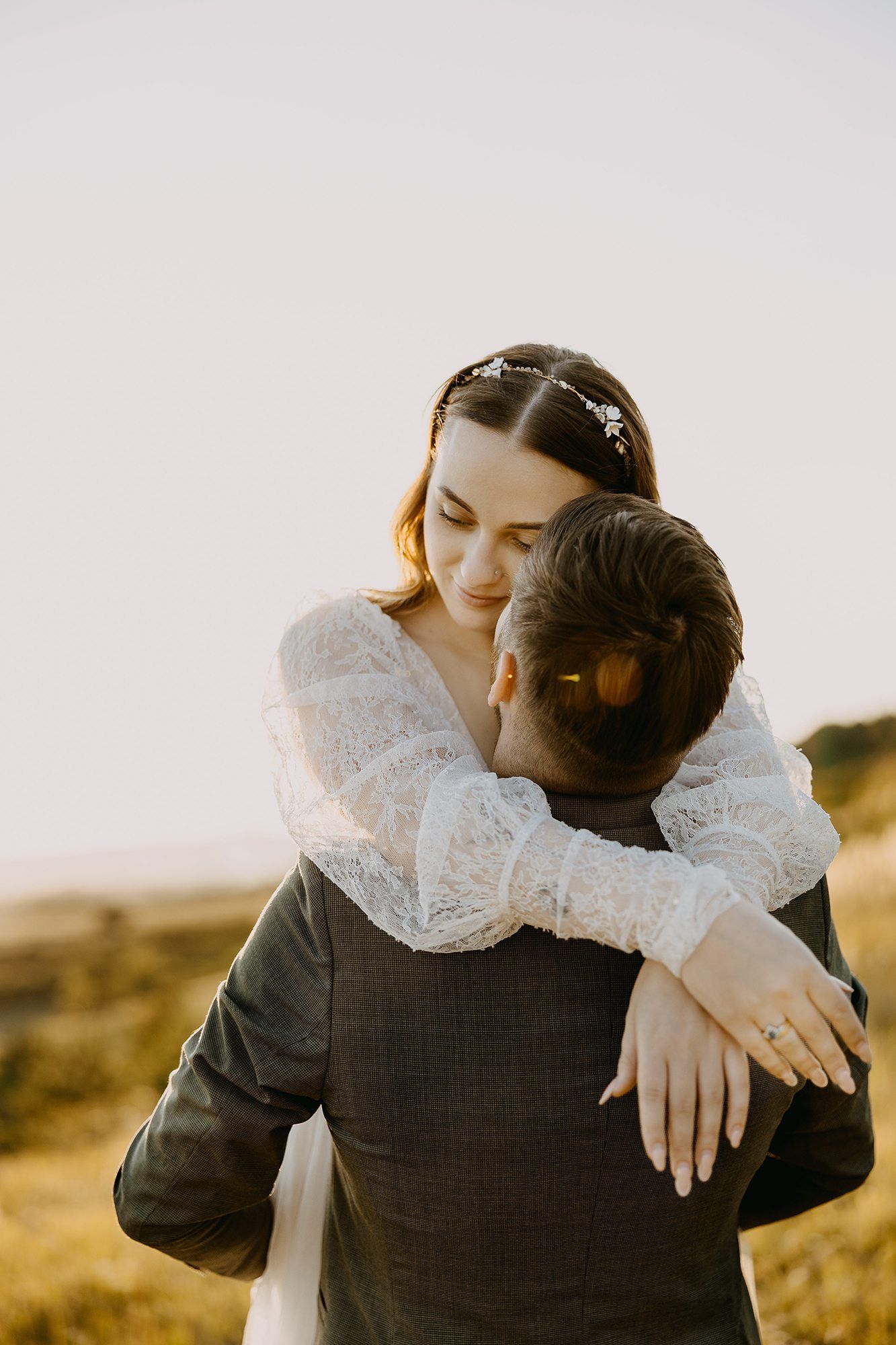 Bride in lace wedding dress hugging groom tenderly during a golden hour outdoor shoot.
