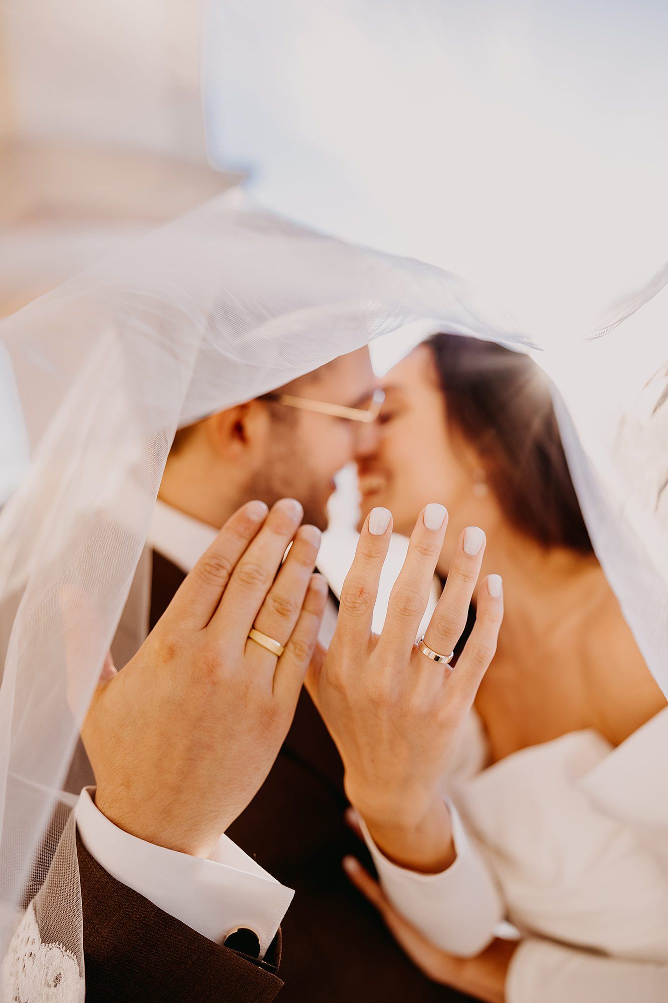 Newlyweds showing gold wedding rings while sharing a kiss under a white veil in soft light.