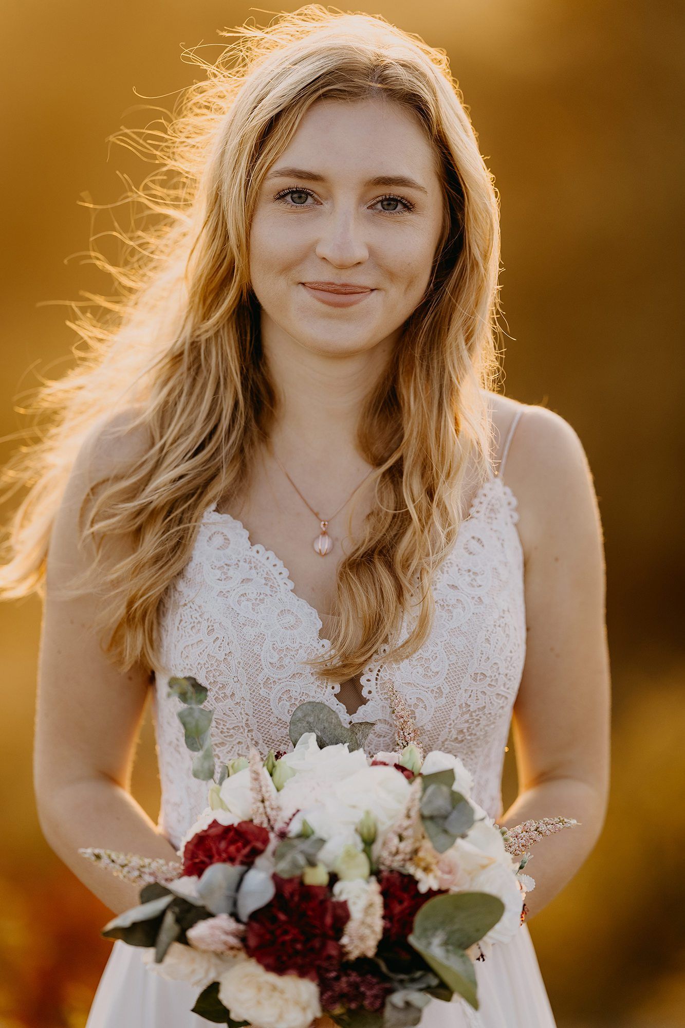 Smiling blonde bride holding a red and white bouquet in warm golden sunset light.