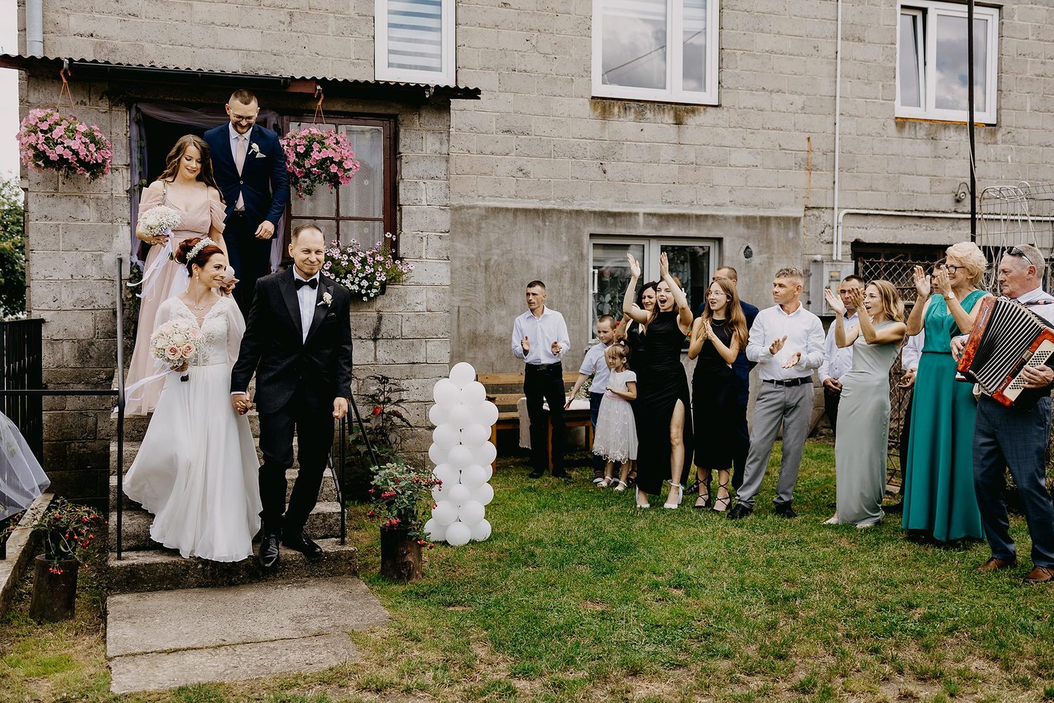 Bride and groom exit home surrounded by wedding guests celebrating outdoors.