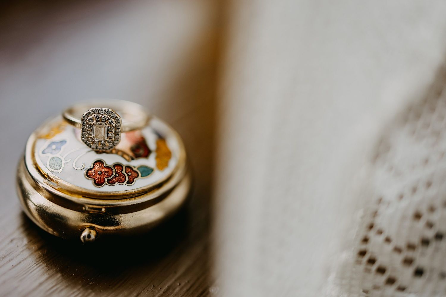 Vintage ornate jewelry box with diamond ring on floral enamel lid on wooden surface.