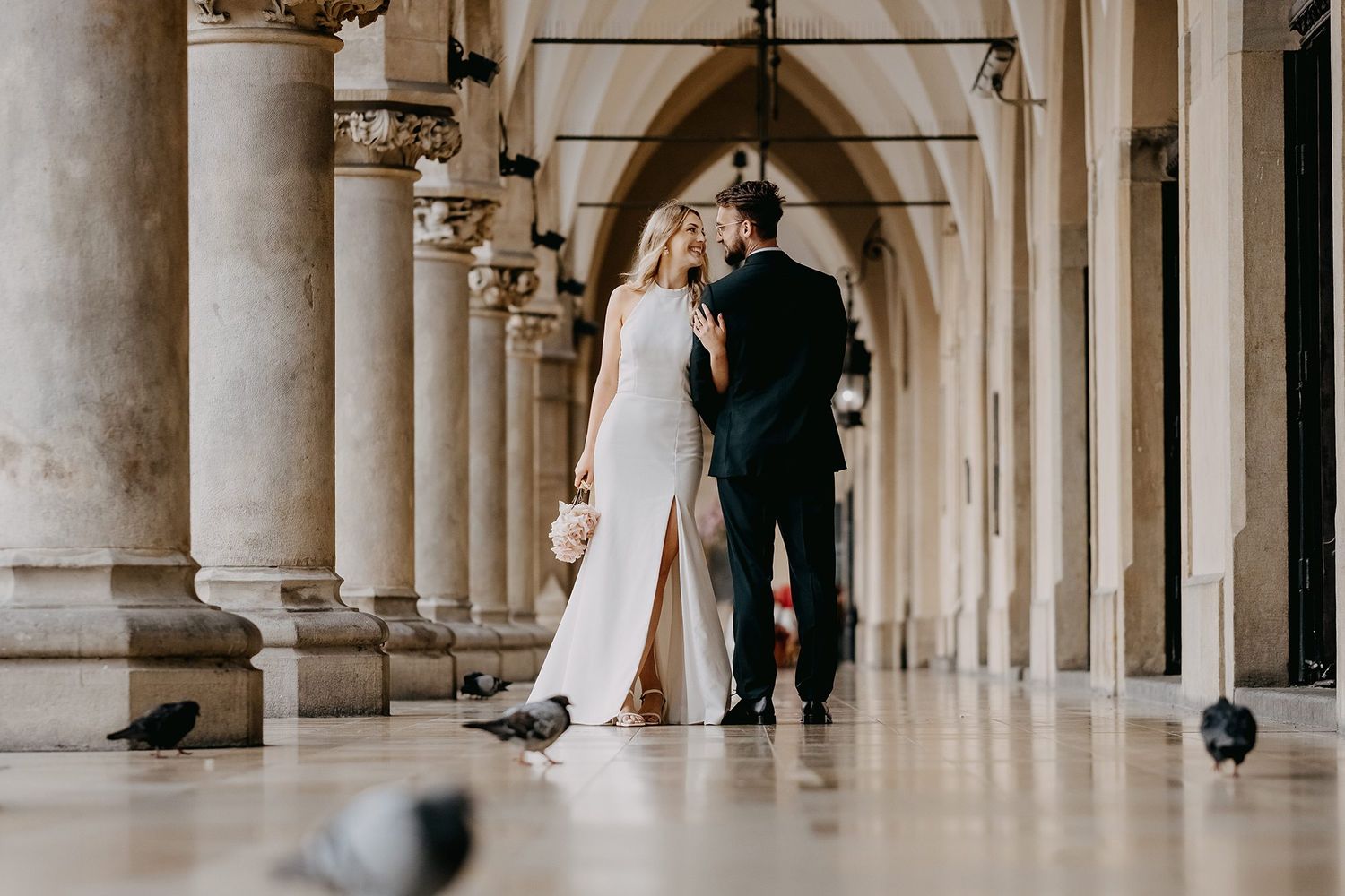 Wedding couple kissing under classical arched colonnade with pigeons nearby.