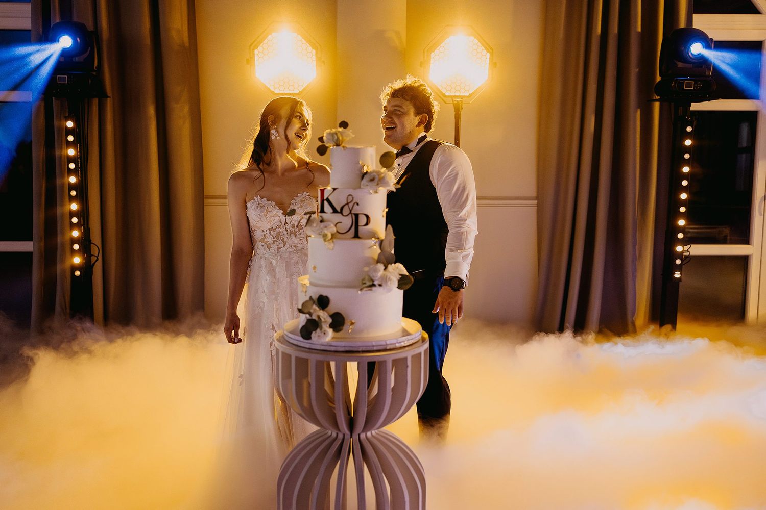 Bride and groom stand beside elegant tiered wedding cake surrounded by dramatic fog effect.