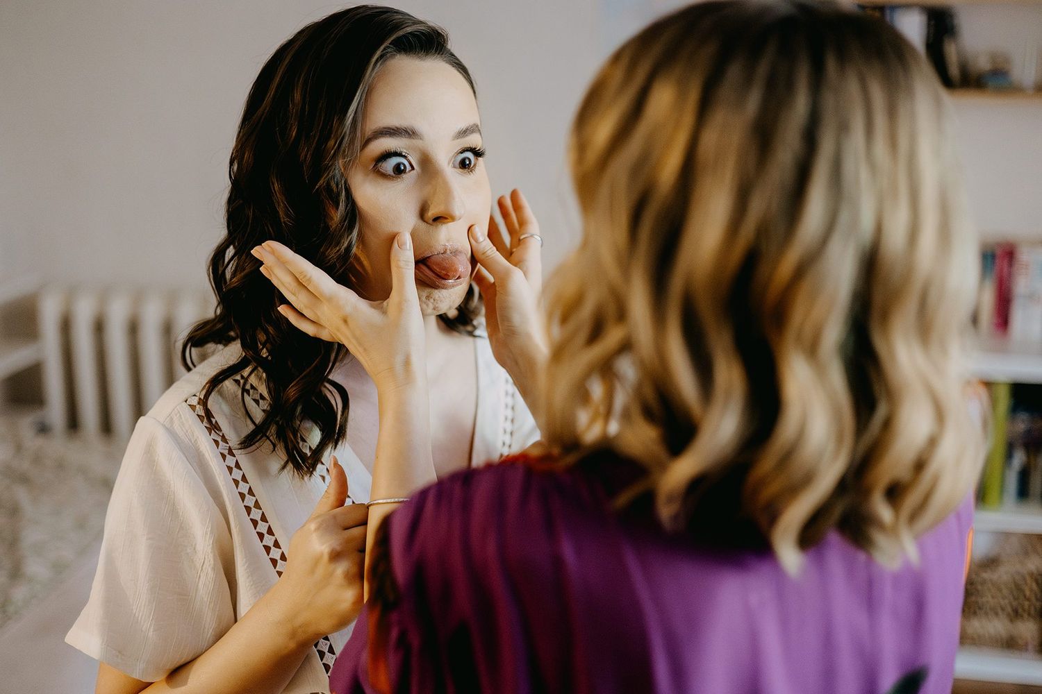 Bridesmaid helping bride with wedding day makeup preparation in a bright room.
