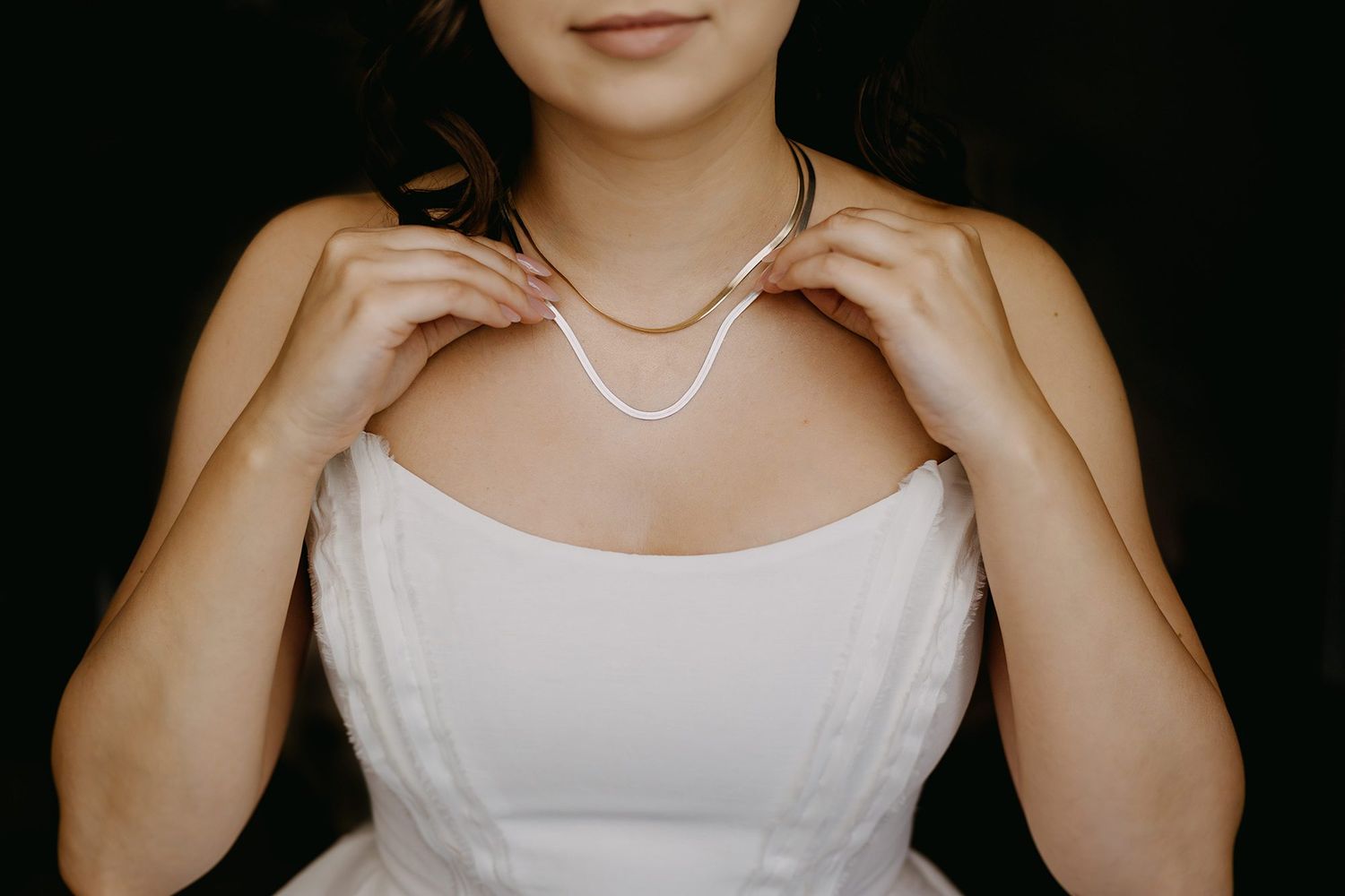 Bride in white strapless gown adjusting pearl necklace against dark background.