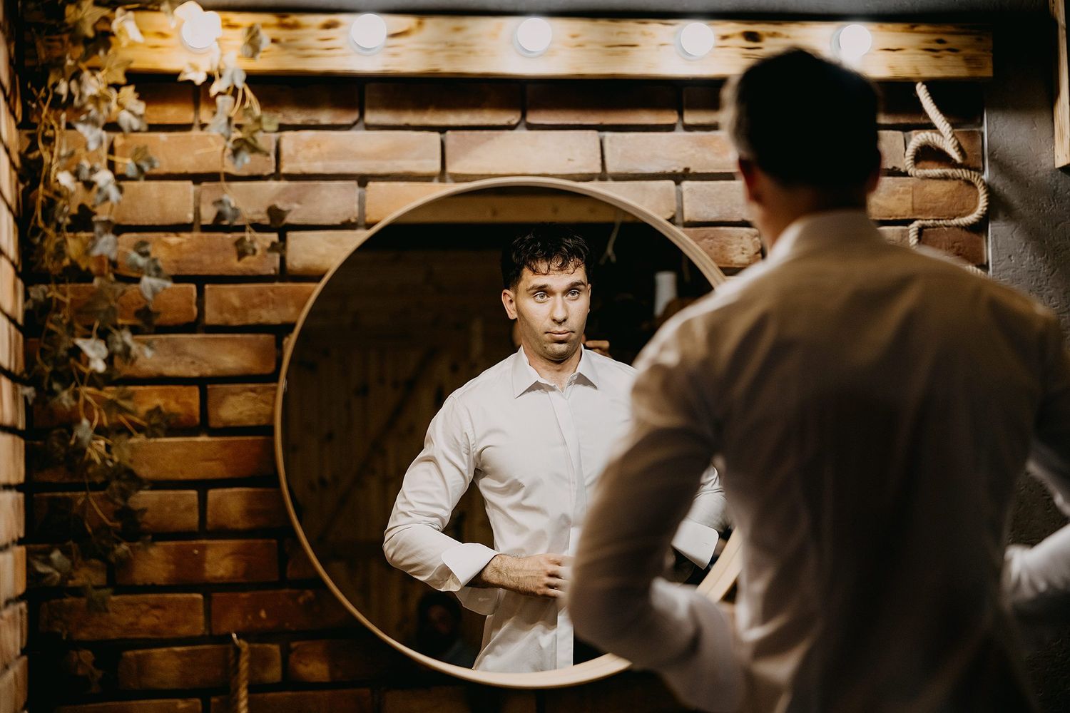Groom in white shirt adjusts his look while gazing into a round mirror on brick wall.