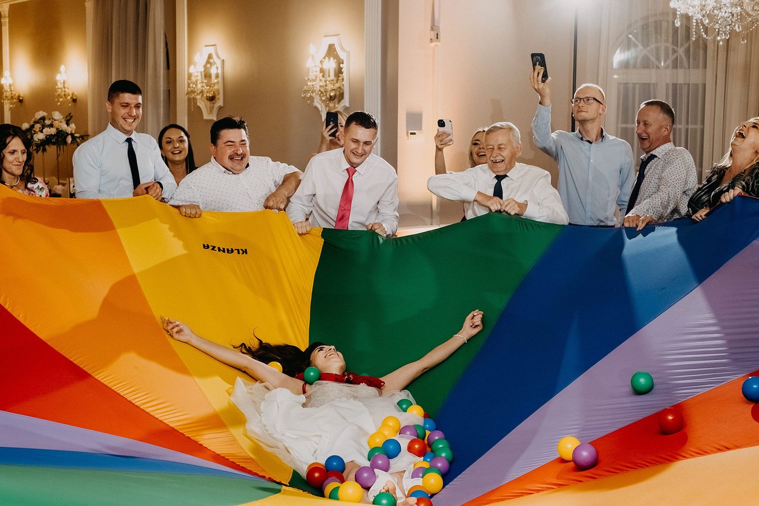 Wedding guests tossing bride on colorful rainbow parachute with balloons at reception.