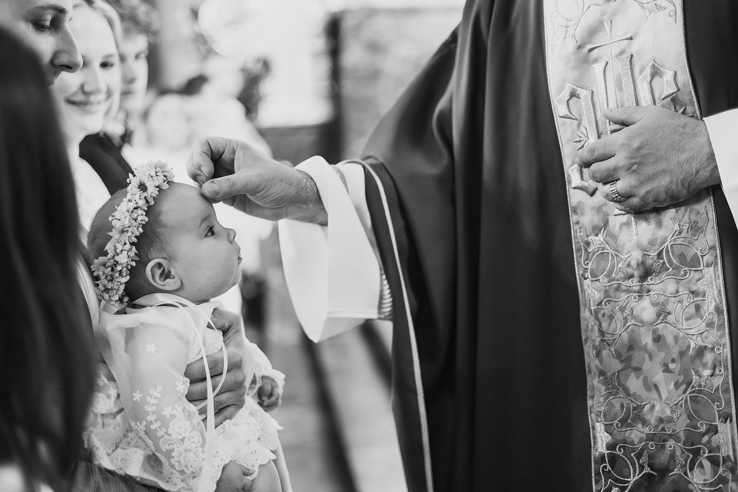 A priest blesses a baby girl in a white dress and floral crown during a baptism ceremony.