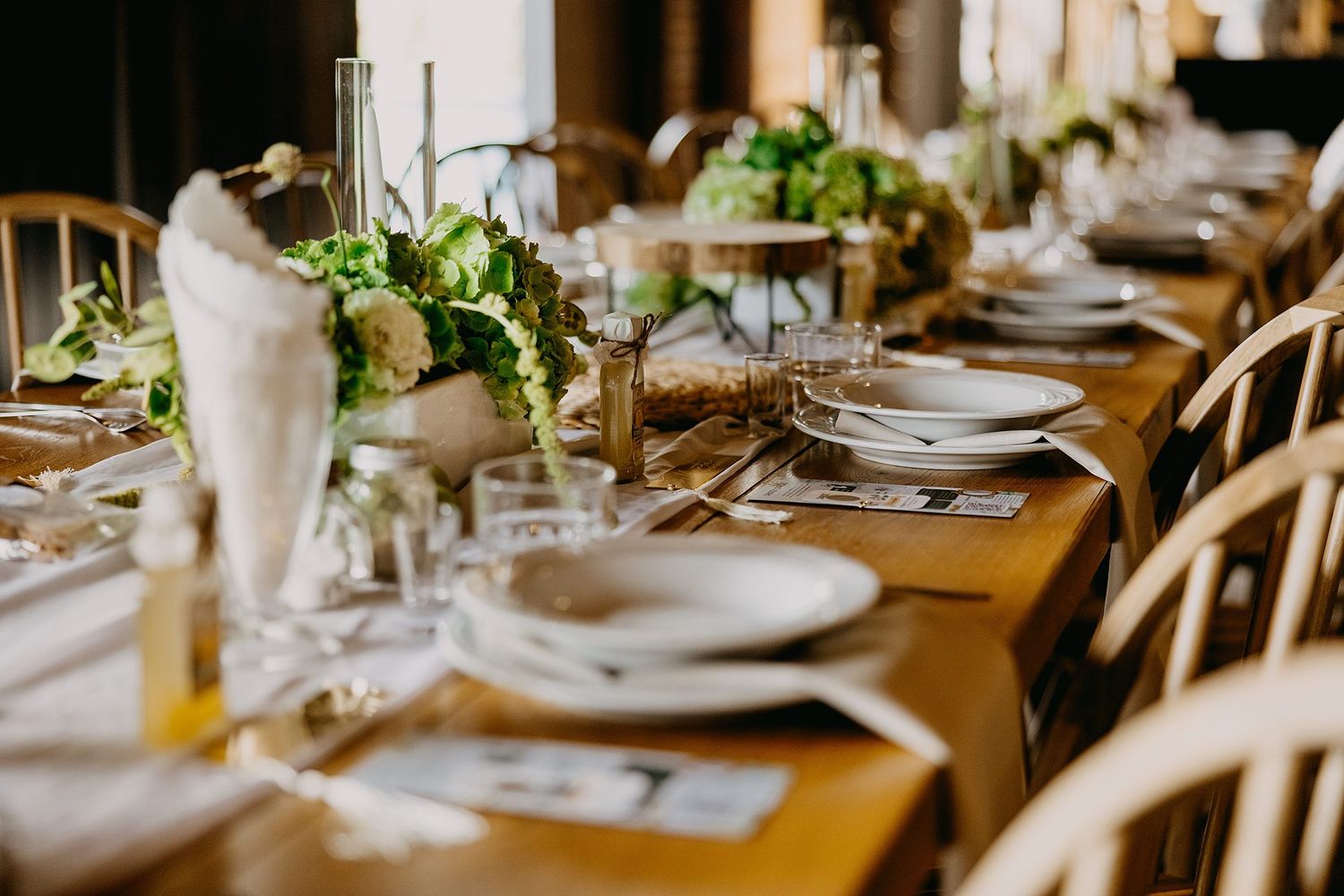 Elegant rustic wedding reception table set with white plates, greenery centerpieces, and wooden chairs in a warm venue.