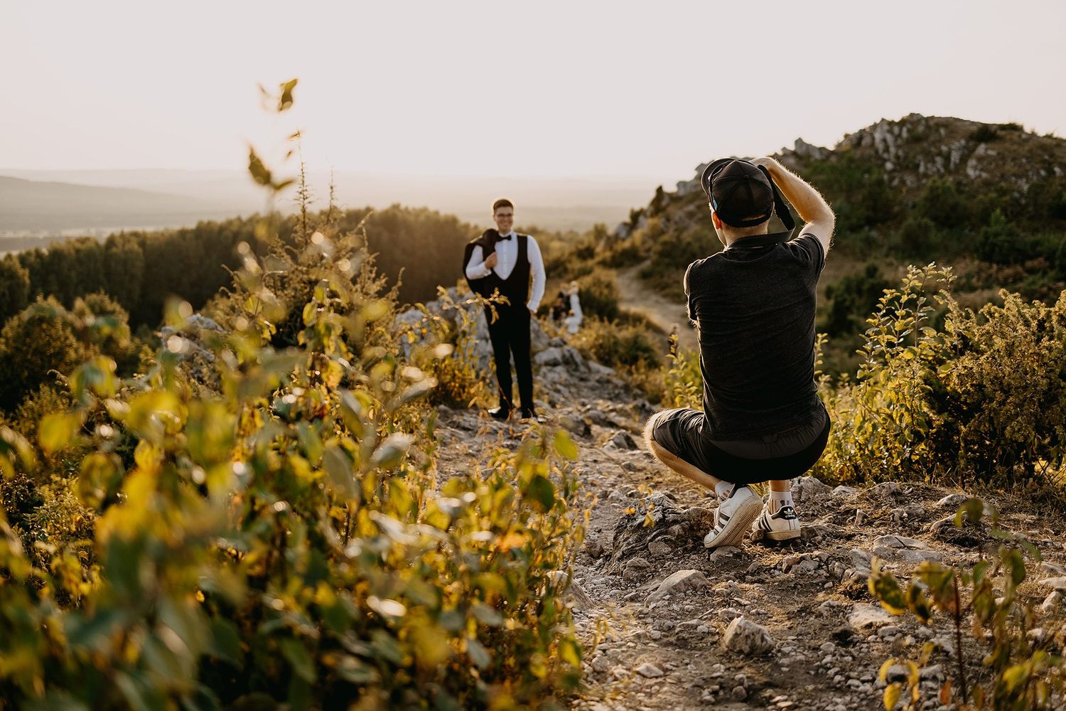 Photographer crouching to capture a man posing on a rocky hillside at golden hour sunset.