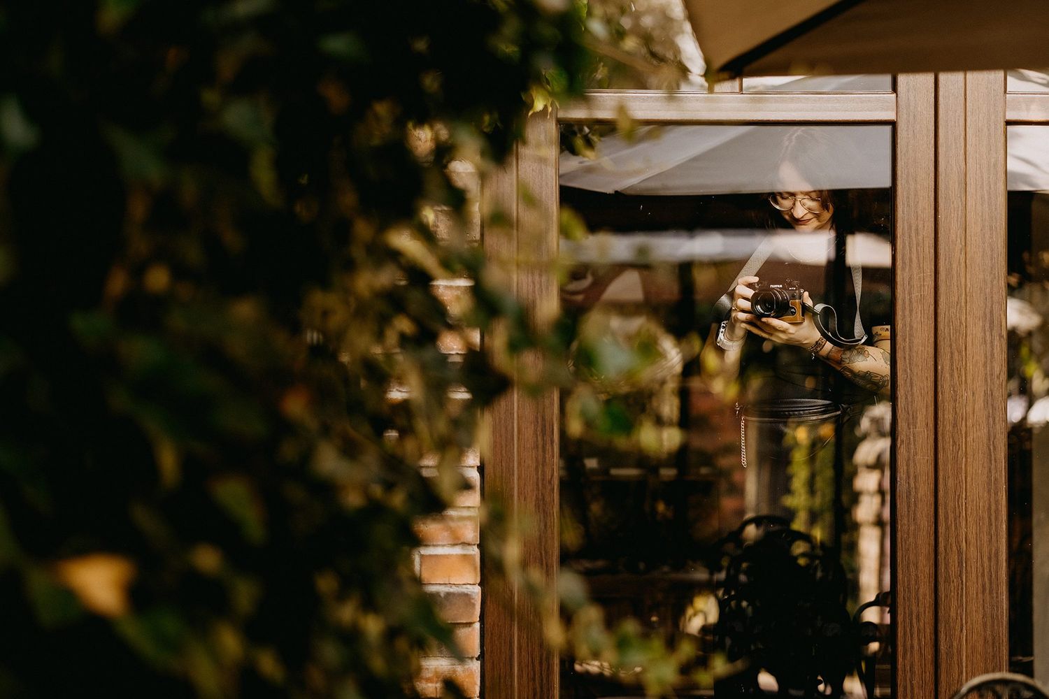 Photographer reflected in glass door surrounded by green foliage outdoors.