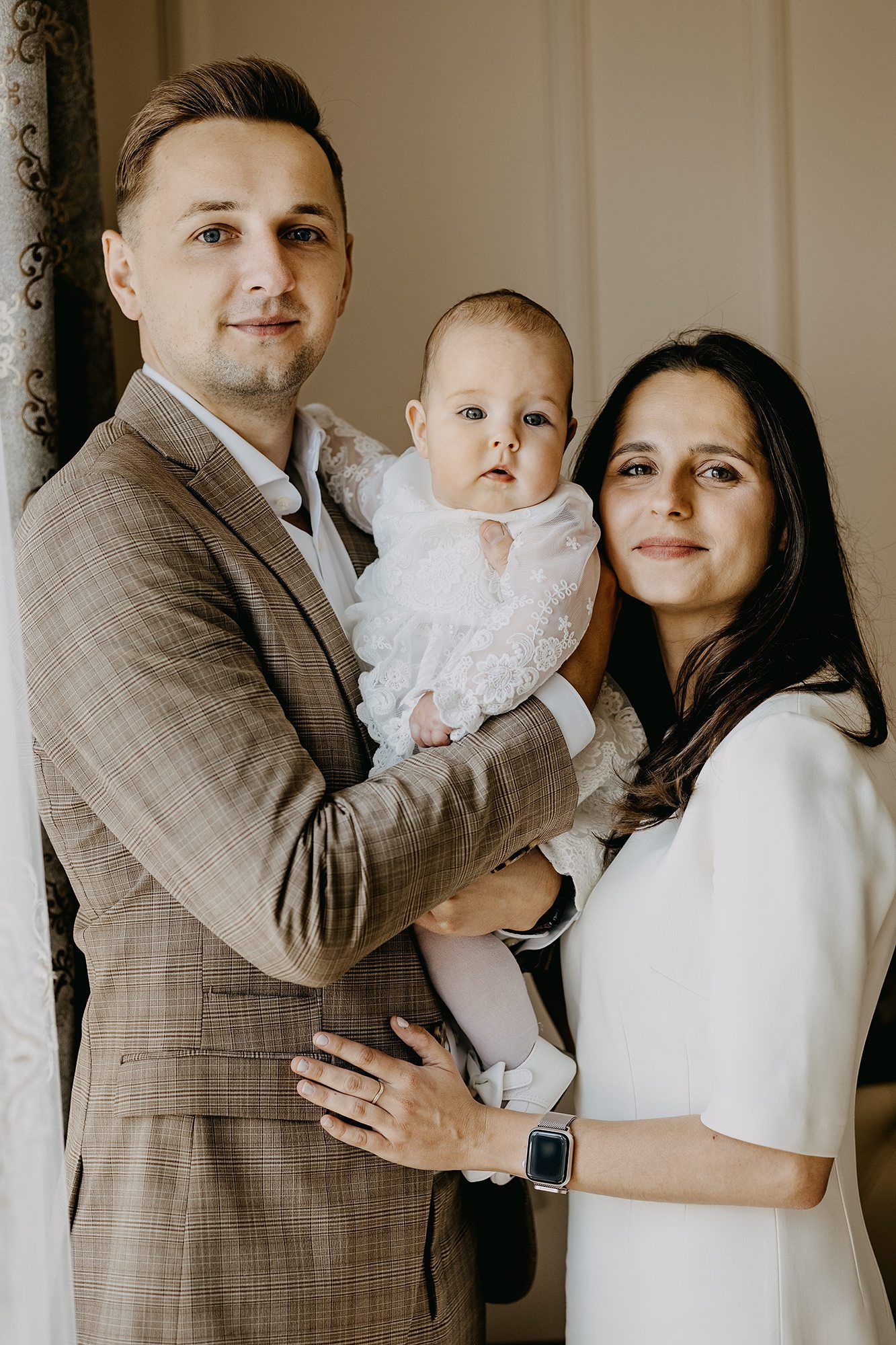 Family portrait of parents in elegant attire holding baby girl in white lace christening dress indoors.