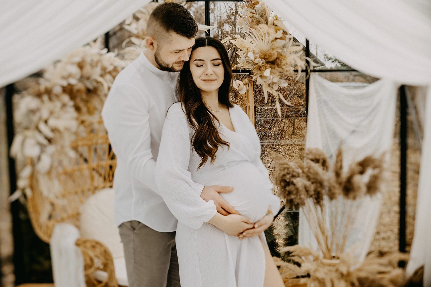 Couple in white outfits embracing during elegant boho maternity photoshoot with pampas grass decor.