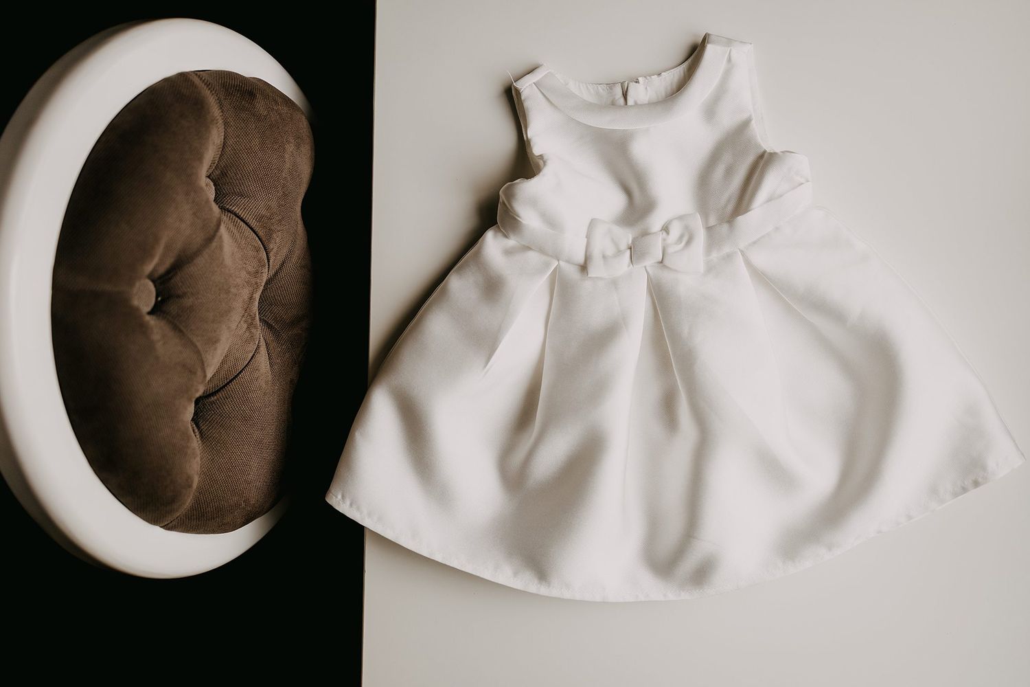 White sleeveless baby girl dress with bow detail displayed flat on white surface next to round velvet cushion.
