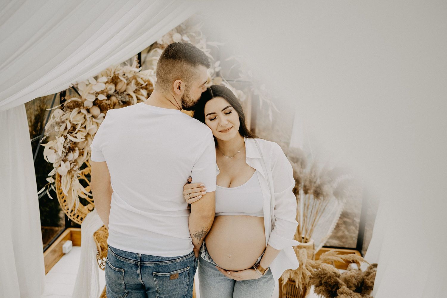 A couple tenderly embraces during a boho-styled maternity photoshoot with dried floral decorations.