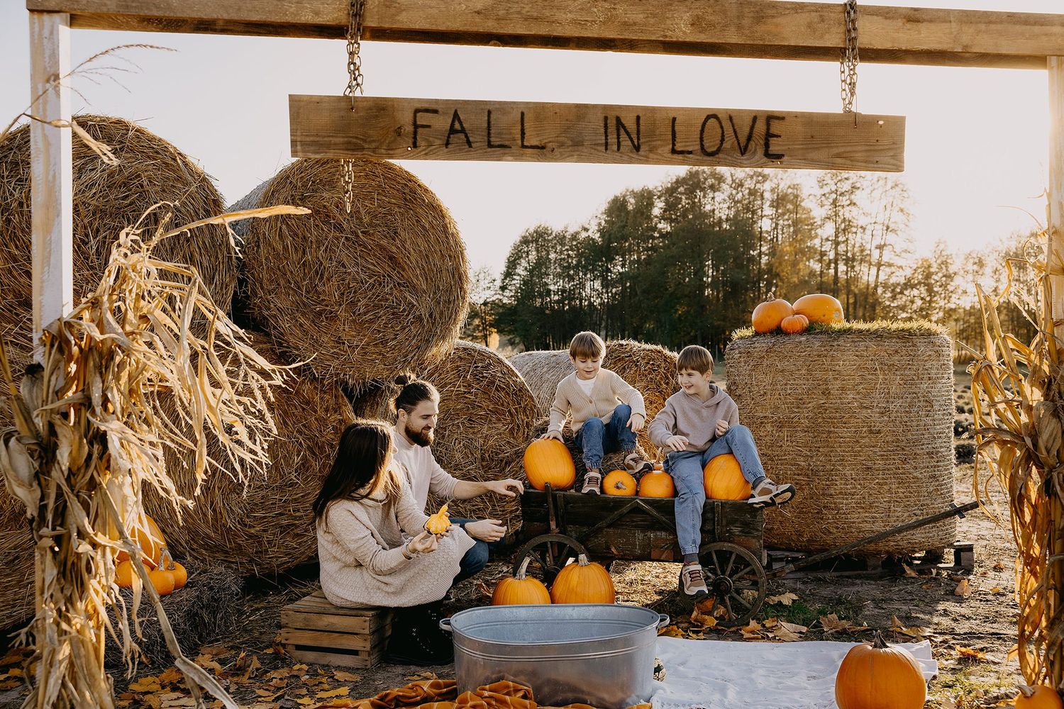 Family carving pumpkins together at a fall harvest display with hay bales and corn stalks.