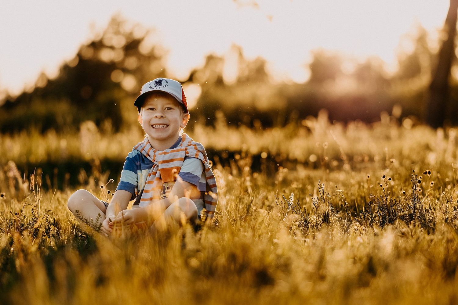 Happy young boy sitting cross-legged in golden sunlit meadow grass, smiling and wearing a striped shirt and cap.