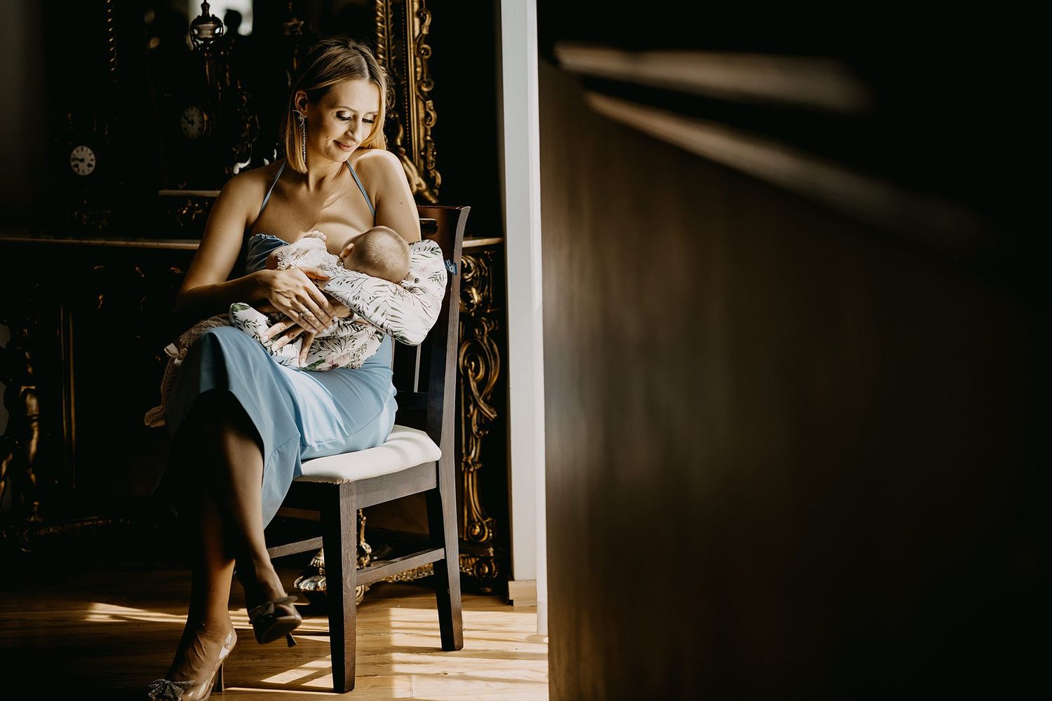 Mother breastfeeding newborn baby while sitting on a chair in warm natural light at home.
