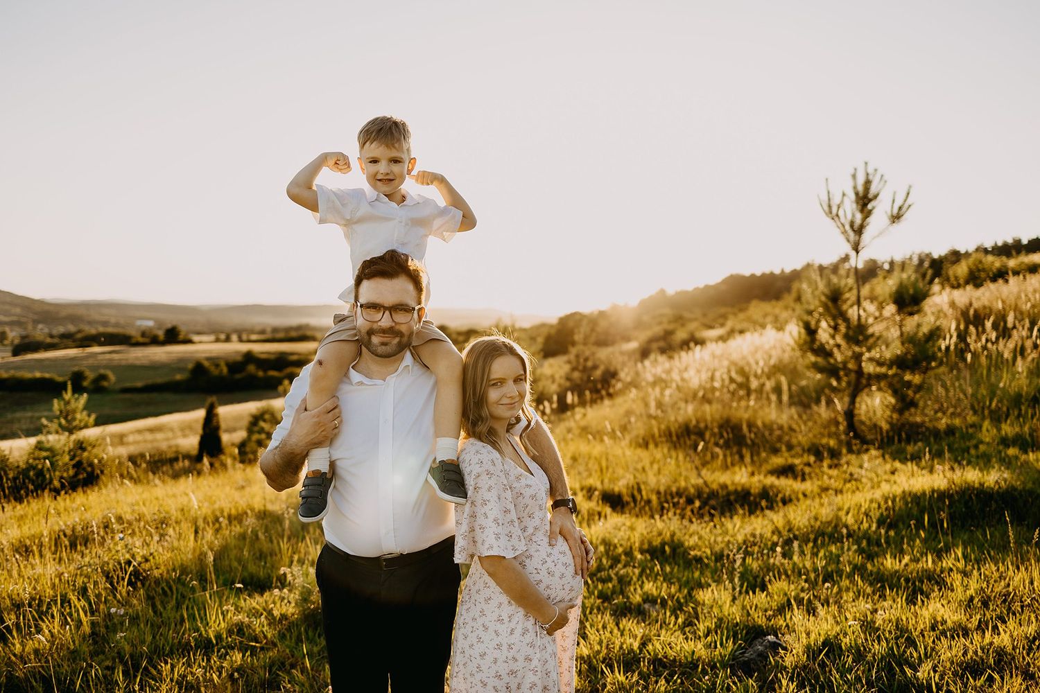 Happy family with pregnant mother, father carrying son on shoulders posing in sunny green hillside.
