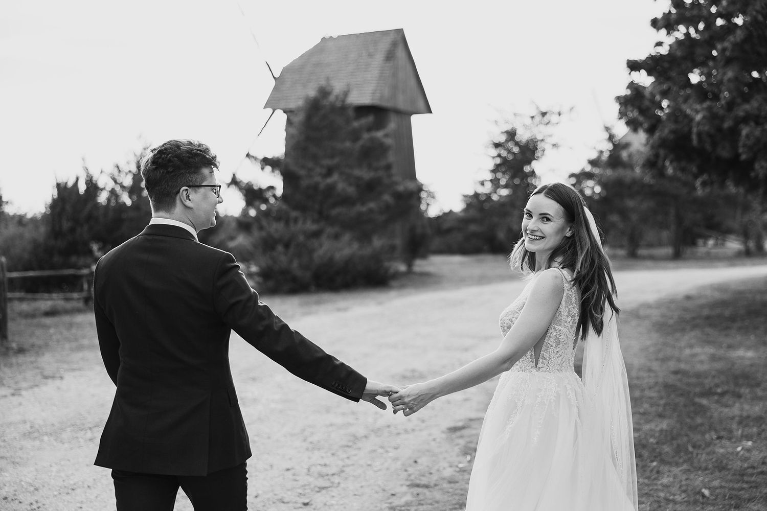 Black and white photo of smiling bride and groom holding hands near rustic wooden chapel outdoors.