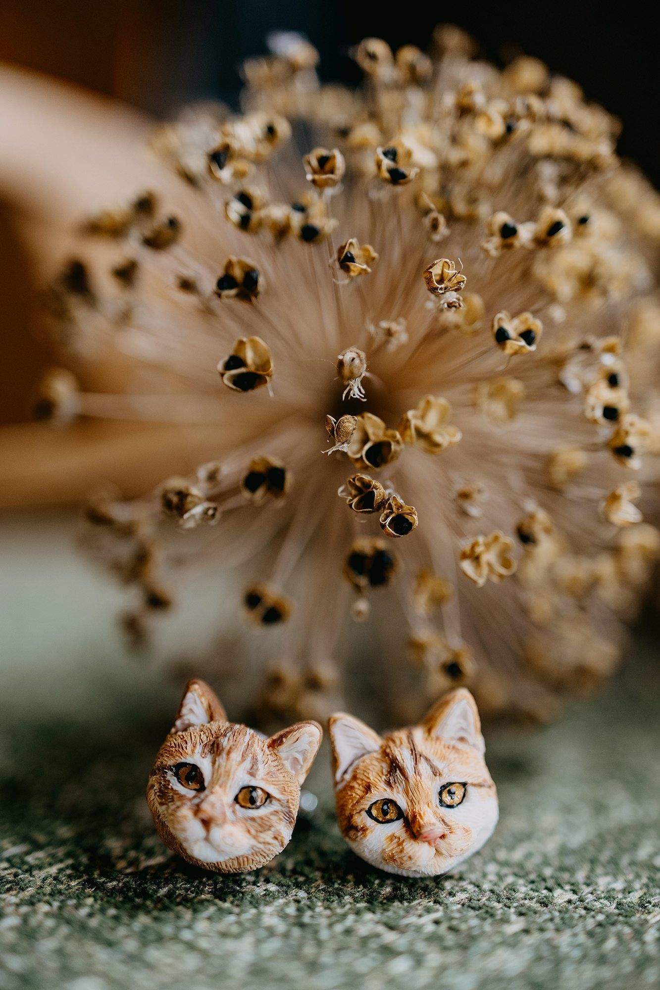 Handcrafted orange tabby cat face stud earrings displayed in front of a dried allium flower.
