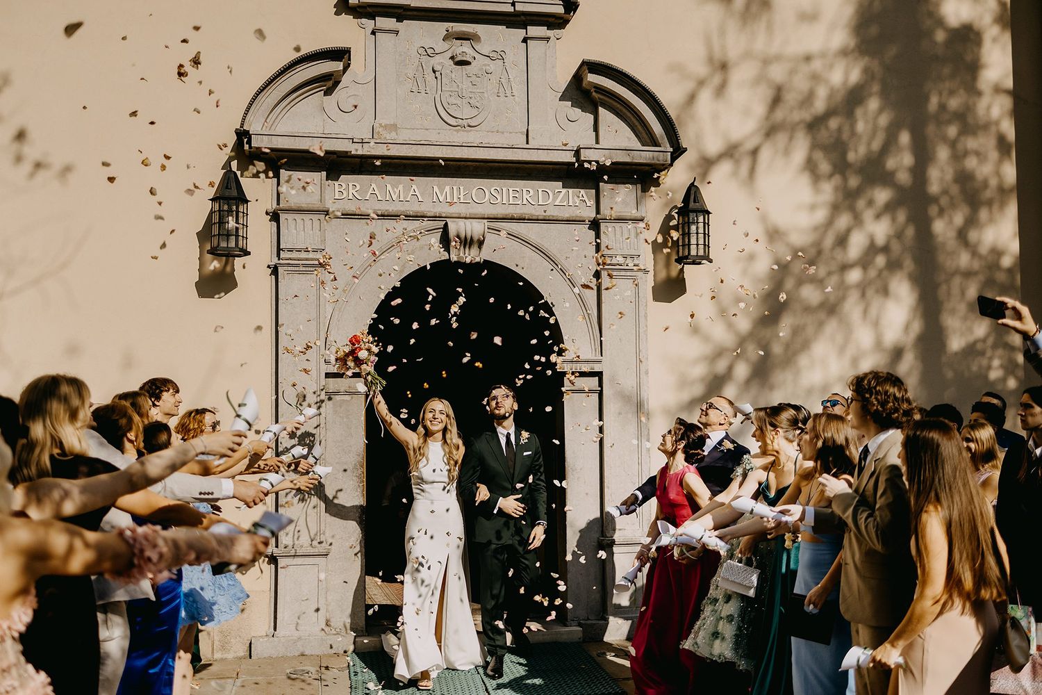 Bride and groom walk through arch as guests shower them with colorful confetti outside.