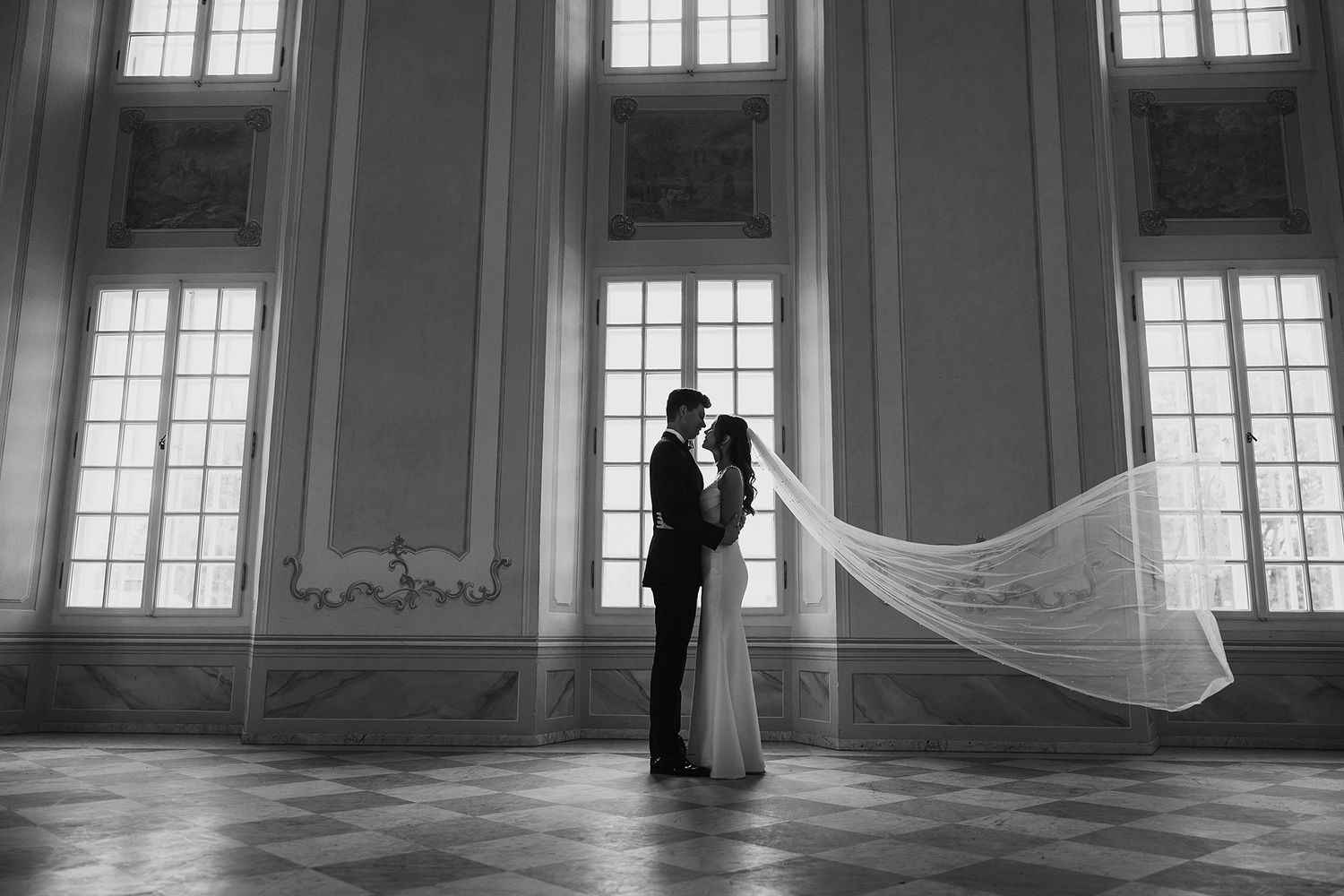 Black and white photo of newlyweds kissing as bride's veil dramatically flows in grand hall.