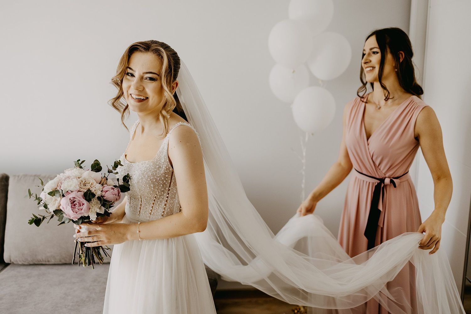 Bride in beaded wedding gown holding pink bouquet while bridesmaid arranges her veil.
