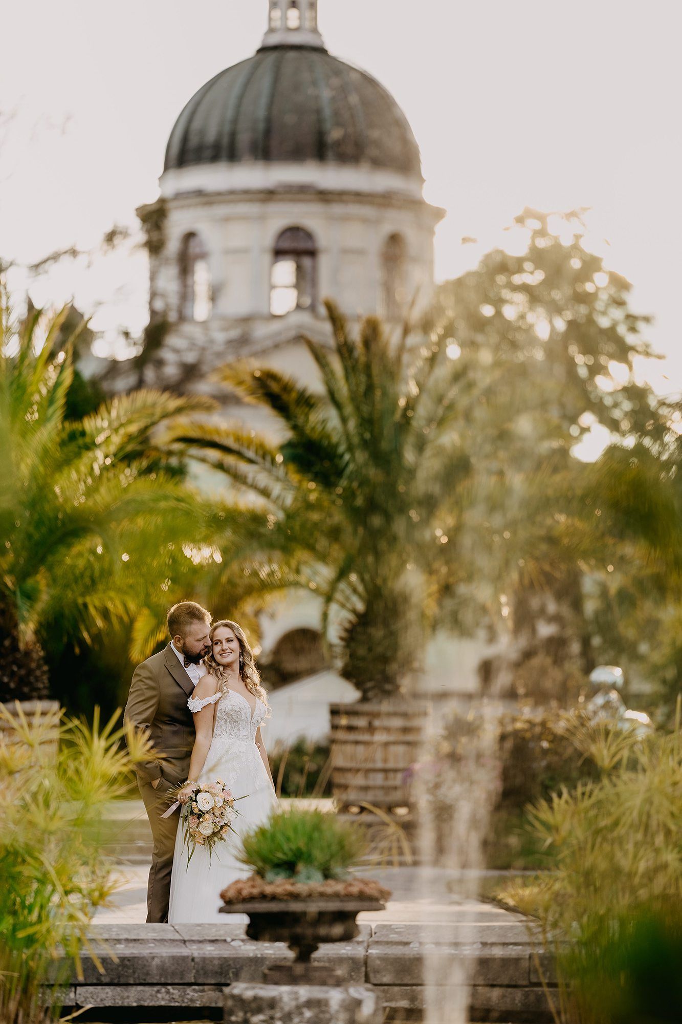 Couple embracing near a fountain in a tropical garden with a domed church tower in soft golden light.