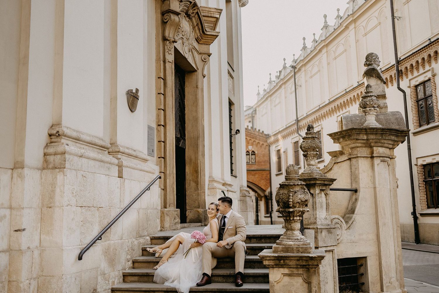 Bride and groom sit together on steps of a classic European historic building in soft daylight.