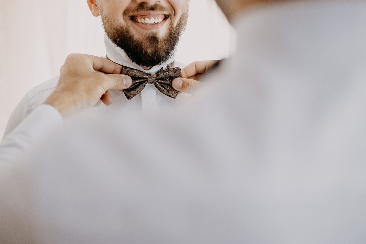 Close-up of smiling bearded groom having patterned bow tie adjusted by groomsman.