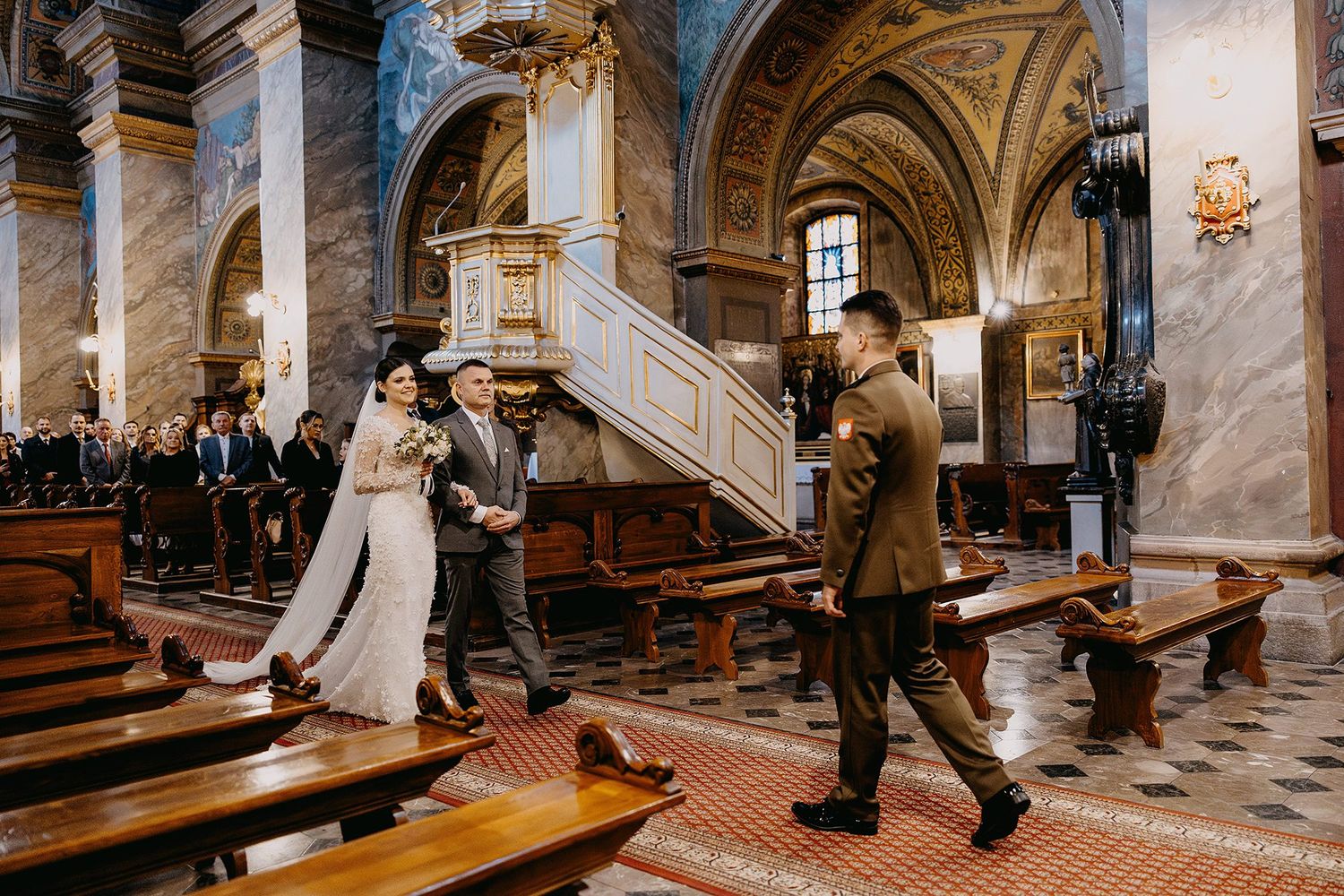 Bride walking down the aisle with escort in an elegant church with wooden pews and marble columns.