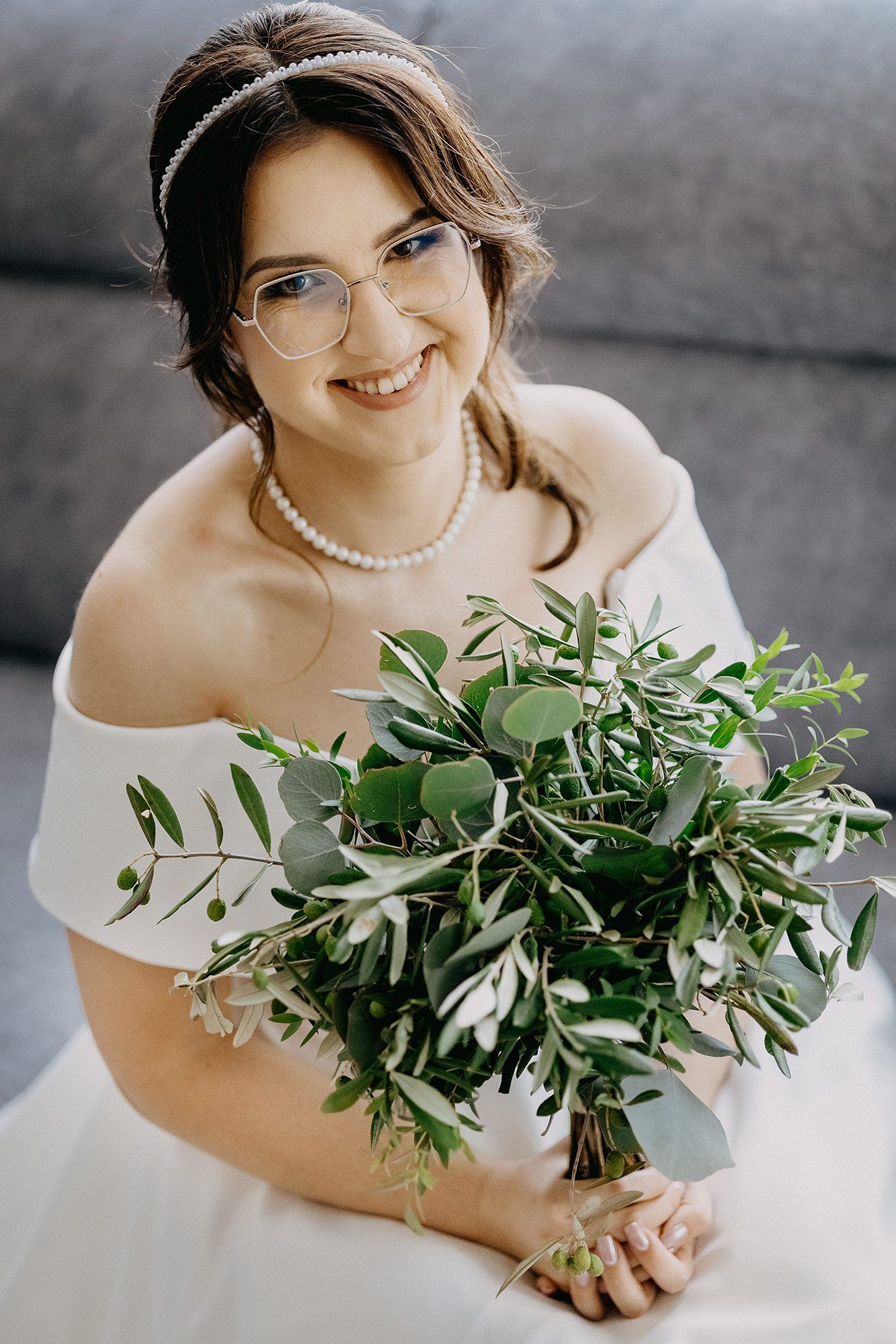 Smiling bride wearing glasses and pearl necklace holding green eucalyptus bouquet in white off-shoulder wedding dress.